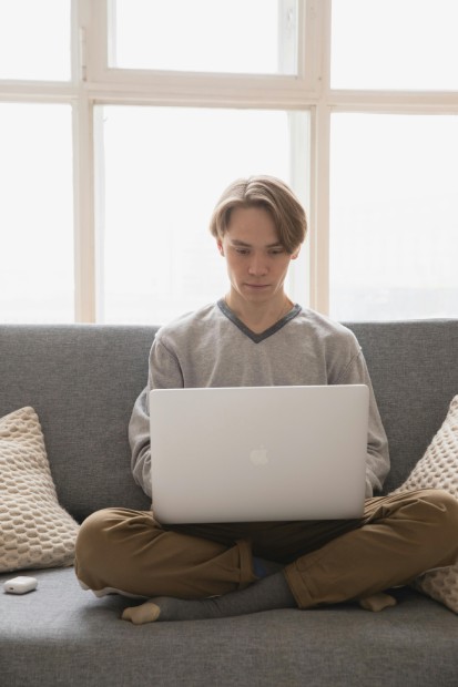 A young person is sitting cross-legged on a grey sofa, using a silver laptop